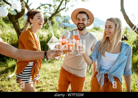 Young and cheerful friends having fun, clinking with wine glasses in the beautifully decorated backyard during a festive meeting or party on a sunny s Stock Photo