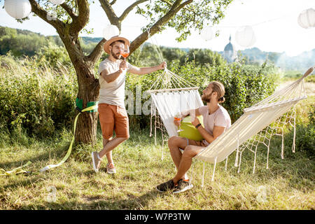 Two male friends having a conversation, drinking wine, relaxing on a hammock in the garden or backyard Stock Photo