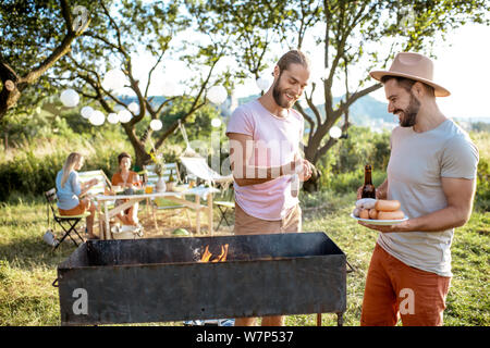 Two male friends cooking food on a barbecue in the beautiful garden during festive lunch with girlfriends on a sunny afternoon Stock Photo