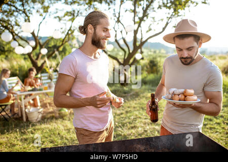 Two male friends cooking food on a barbecue in the beautiful garden during festive lunch with girlfriends on a sunny afternoon Stock Photo