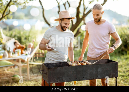 Two male friends cooking food on a barbecue in the beautiful garden during festive lunch with girlfriends on a sunny afternoon Stock Photo