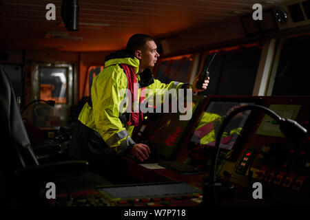 Navigator. pilot, captain as part of ship crew performing daily duties with VHF radio, binoculars, logbook, standing nearby to ECDIS and radar station Stock Photo