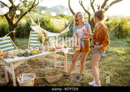 Two girlfriends talking together on a picnic in the decorated garden with table full of healthy food on a summer evening Stock Photo