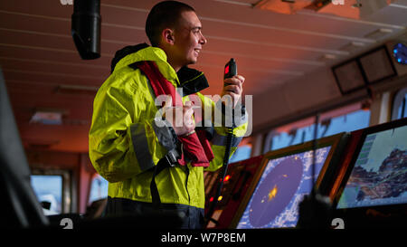 Navigator. pilot, captain as part of ship crew performing daily duties with VHF radio, binoculars, logbook, standing nearby to ECDIS and radar station Stock Photo