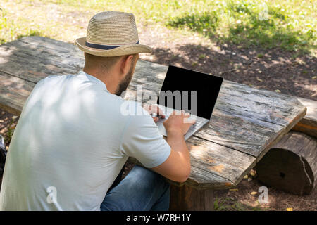 Man or guy working on a computer with a blank screen for copy space, sitting on a bench in the summer outdoors in a park. rear view from the shoulder Stock Photo