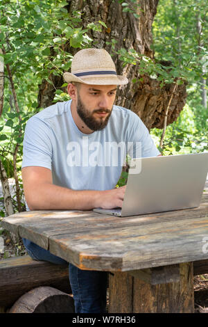Young, bearded guy in a hat sits on a bench in the summer on the street, using a laptop, looking at the screen, a man working, studying or surfing the Stock Photo