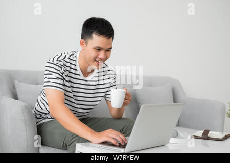 Young man concentrating on laptop computer screen, sitting at desk in living room, holding mug.? Stock Photo