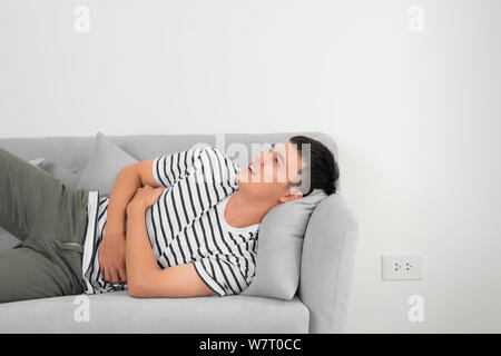 Terrible stomachache. Frustrated handsome young man hugging his belly and keeping eyes closed while lying on the couch at home Stock Photo
