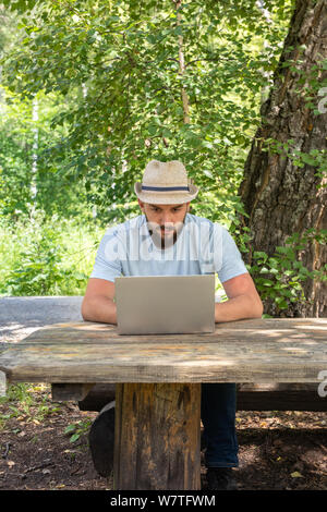 Young attractive 23 year old man, guy with a beard looks at the laptop screen, sitting on a bench in a park outdoors. Searches for a girlfriend on the Stock Photo
