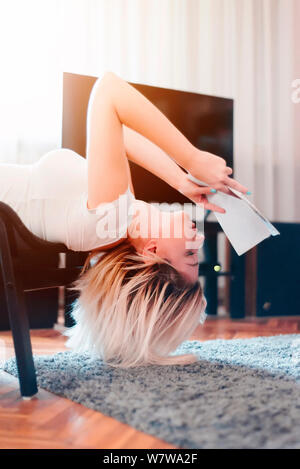 Young woman reading a magazine on chair upside down in living room Stock Photo