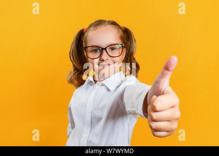 Portrait of a cute attractive little girl in glasses. Child shows thumb up on a yellow background. Stock Photo