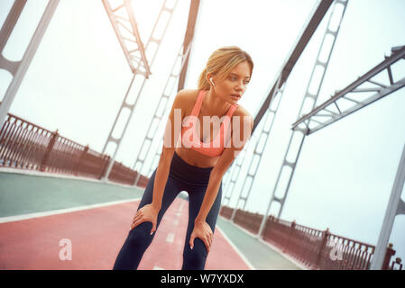 Feeling exhausted. Portrait of young tired blonde woman in sports clothing relaxing after jogging and looking aside while standing on the bridge outdoors. Sport concept. Healthy lifestyle. Taking a break Stock Photo