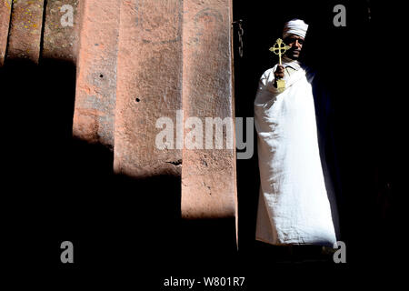 Priest in traditional white robes and turban, holding a gold cross. Bet Giyorgis Church, Lalibela. UNESCO World Heritage Site. Ethiopia, December 2014. Stock Photo