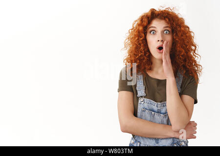 Shocked and speechless cute modern redhead curly-haired girl, gasping amazed, drop jaw fascinated and impressed, stare camera, cover opened mouth with Stock Photo