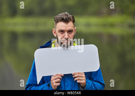 Upset young man holds a blank poster with a mockup for your advertisement on the background of nature. Stock Photo