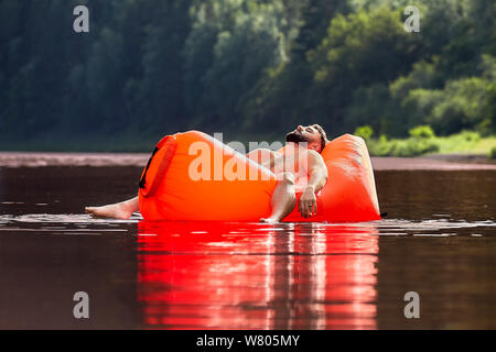 An orange inflatable lounger is drifting along the river, a handsome bearded young man is sleeping in a lounger, resting on nature during vacation. Stock Photo