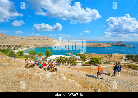 Tourists ride donkeys up the hill towards the Lindos Acropolis with the Bay of Lindos in view on the island of Rhodes, Greece Stock Photo