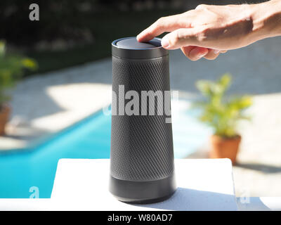 Cropped image of a man using a portable speaker with voice assistant and touch technology near the outdoor swimming pool. Stock Photo