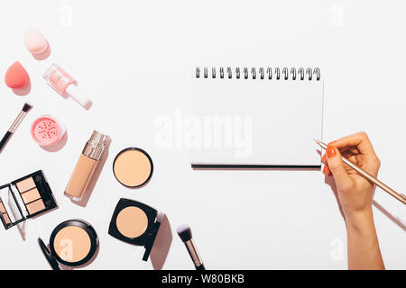 Female's hand writing in blank blank notebook next to makeup products and accessories on white background, flat lay. Stock Photo