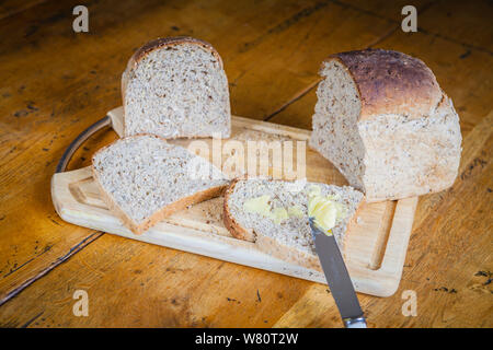 sliced granary bread with knife spreading butter. on a wooden bread board on wooden farmhouse table. Stock Photo