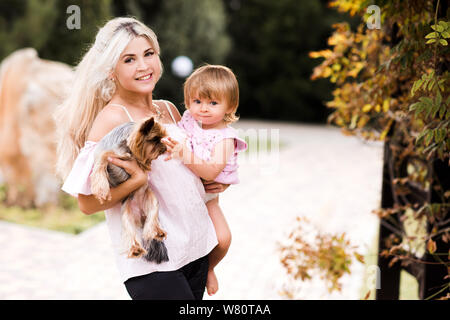 Happy mother holding baby 1 year old daughter and small pet dog outdoors over nature background in park. Looking at camera. Motherhood. Maternity. Stock Photo