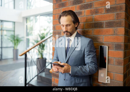 Grey-haired businessman using smartphone standing near elevator Stock Photo