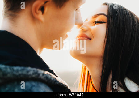 Side view close up portrait of charming european couple trying to kiss with closed eyes smiling against sunset while dating outside against sunset. Stock Photo