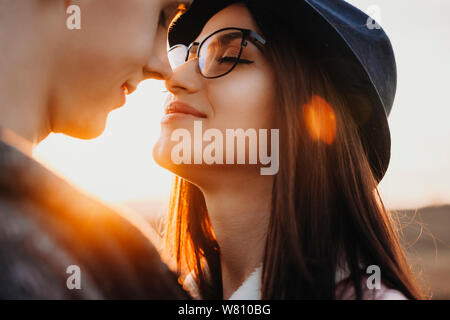 Close up portrait of a beautiful woman with dark hair wearing hat and glasses smiling and trying to kiss her boyfriend against sunset while dating out Stock Photo