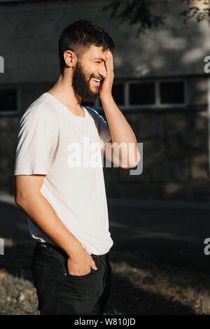 Portrait of a young bearded caucasian male laughing with closed eyes and touching his face with a hand while walking through city. Stock Photo