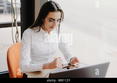 Cute adult business woman sitting operating on smartphone while sitting in her office working at laptop near a window. Stock Photo