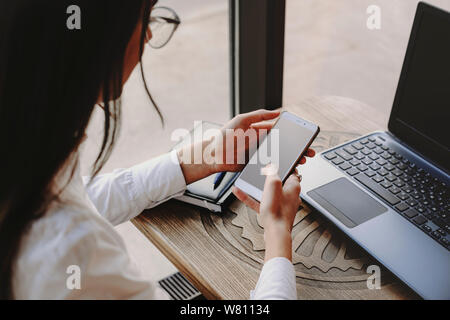 Female hands using a smartphone for operating on internet transaction and laptop sitting at a table in coffee shop. Stock Photo