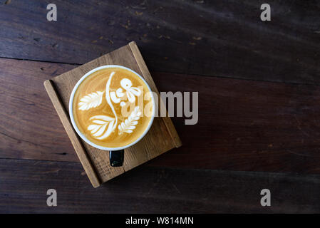 Top view a cup of hot coffee cappuccino with cute signature rabbit, flower and leaf latte art pattern in black ceramic cup on rough wooden table. Stock Photo