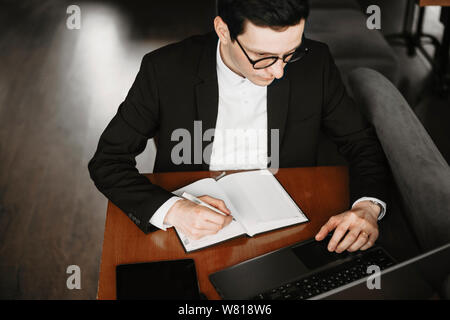 Upper view of a male hand writing in a notebook and operating on a laptop while sitting on a table. Stock Photo