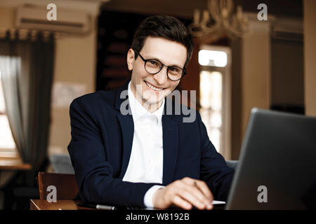 Portrait of a attractive confident businessman dressed in suit and wearing glasses looking at camera smiling while working at his laptop in his loft o Stock Photo