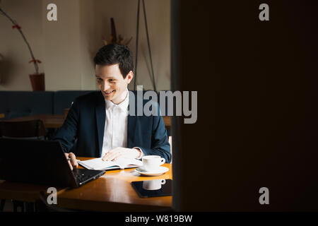 Young stylish manager doing paperwork while operating on his laptop smiling while sitting in a restaurant. Stock Photo