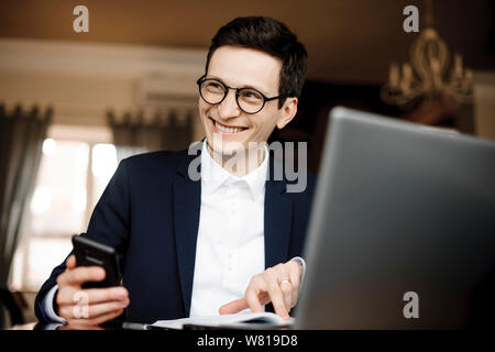 Portrait of a confident handsome businessman working while holding a smartphone and pointing with finger in his notebook looking away laughing dressed Stock Photo