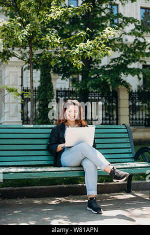 Full length portrait of a handsome plus size woman sitting on a bench with a laptop on her legs looking at the laptop smiling against a building. Stock Photo