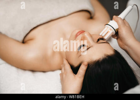 Upper view of a charming young woman doing mesotherapy on her face in clinic salon while leaning on a bed with closed eyes. Stock Photo