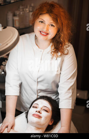 Portrait of a lovely cosmetologist with red hair looking at camera smiling while finishing to apply a white organic mask to a female client in a spa s Stock Photo