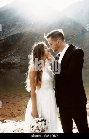 Side view portrait of a cute young caucasian couple looking to each other smiling before kissing against lake and mountain in the morning. Stock Photo