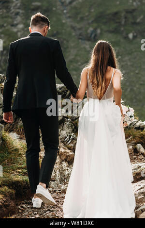 Back view of a unrecognizable caucasian young groom and bride holding hands and walking in mountains. Stock Photo