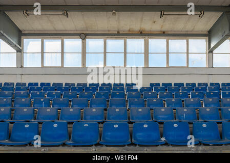 Front view row of blue plastic seats on rough concrete step floor in old vintage dirty concrete structural stadium without people. Stock Photo