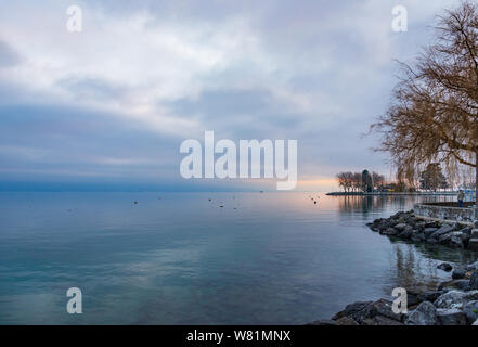 Beautiful tranquil natural walkway and promenade along lakeside of lake Geneva and background of misty, cloudy and twilight sky over water. Stock Photo