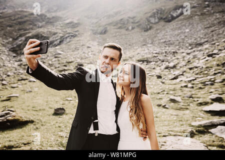 Beautiful young caucasian bride and groom doing a selfie on the smartphone in front of mountains in their wedding day while groom is showing tongue. Stock Photo