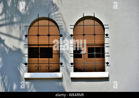Pair of arched Arabic style windows with metal grills on the yellow stucco exterior of Singapore house with shadow of palm tree Stock Photo