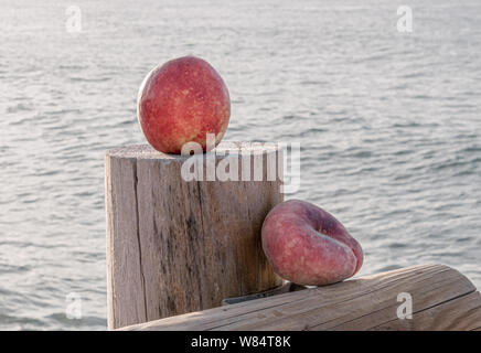 Delicious dessert with the calm sea in the background - A beautiful red apricot accompanied by a flat red peach on a wooden dock Stock Photo