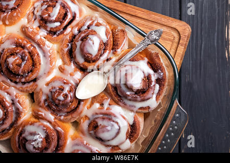Delicious cinnamon buns in a baking tray glazed with cream cheese frosting, standing on the black wooden table, top view Stock Photo