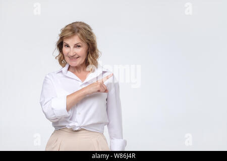 Beautiful old woman in white shirt is pointing to the side, looking at camera and smiling Stock Photo