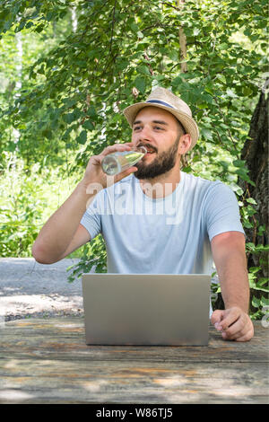 Heat in the street, thirsty. A young attractive man with a beard of Caucasian appearance drinks clean water from a bottle. Sitting in the park while a Stock Photo