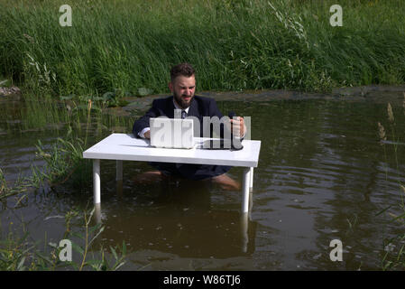 Frustrated, displeased young bearded man, guy. Trying to focus on work. tired of office work. Sitting at a table standing in a swamp in the water Stock Photo
